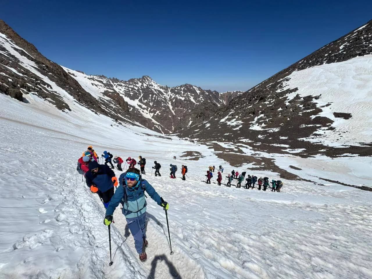 Des randonneurs montent en file sur une pente enneigée, entre des versants rocheux, sous un ciel bleu